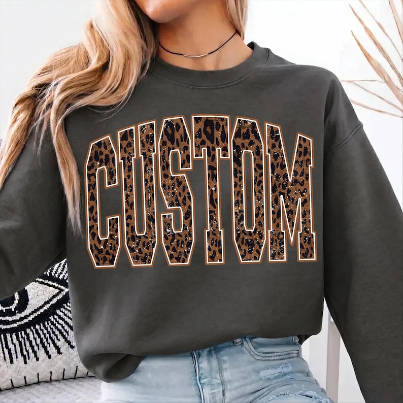 May include: A dark gray crewneck sweatshirt featuring the word CUSTOM printed in large, block-style letters across the chest. The letters have a brown and black leopard print pattern with a thin white outline, creating a bold graphic design.