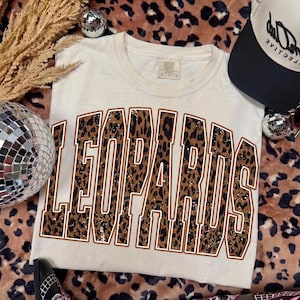 May include: A cream-colored short-sleeve t-shirt featuring the word LEOPARDS in large, block-style letters. The letters have a brown and black leopard print pattern with a white outline. The shirt has a crew neckline and a tag at the inner collar.