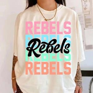 May include: Cream-colored t-shirt with the word "REBELS" in a stacked design. The word is in pink, blue, black, and teal. The shirt has short sleeves and a relaxed fit.