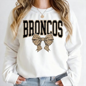 May include: White sweatshirt featuring the word "BRONCOS" in black block letters with a leopard print fill. A leopard print bow is centered below the word. The sweatshirt is a crew neck style.