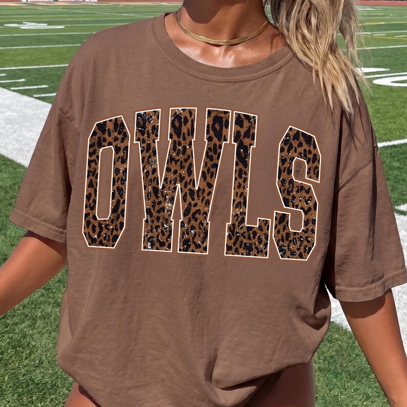 May include: A brown short-sleeve t-shirt featuring the word OWLS printed in large, block-style letters across the chest. The letters have a brown and black leopard print pattern with a thin white outline, creating a bold graphic design.