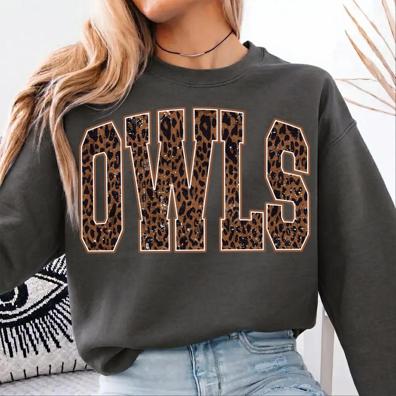 May include: A dark gray crewneck sweatshirt featuring the word OWLS in large, block-style letters. The letters have a brown and black leopard print pattern with a thin white outline, centered on the front of the garment.