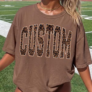 May include: A brown short-sleeved t-shirt featuring the word CUSTOM printed in large, block-style letters across the chest. The letters have a brown and black leopard print pattern with a thin white outline, creating a bold graphic design.