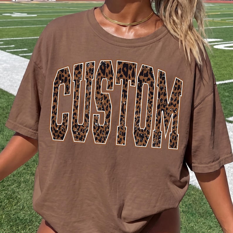 May include: A brown short-sleeved t-shirt featuring the word CUSTOM printed in large, block-style letters across the chest. The letters have a brown and black leopard print pattern with a thin white outline, creating a bold graphic design.