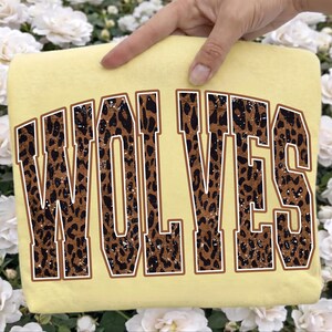 K&ouml;nnte beinhalten: Ein blassgelbes T-Shirt mit dem Wort WOLVES in gro&szlig;en Blockbuchstaben im College-Stil. Die Buchstaben haben ein braun-schwarzes Leopardenmuster mit einer abgenutzten, gesprenkelten Textur und einer d&uuml;nnen wei&szlig;en Umrandung um jedes Zeichen.