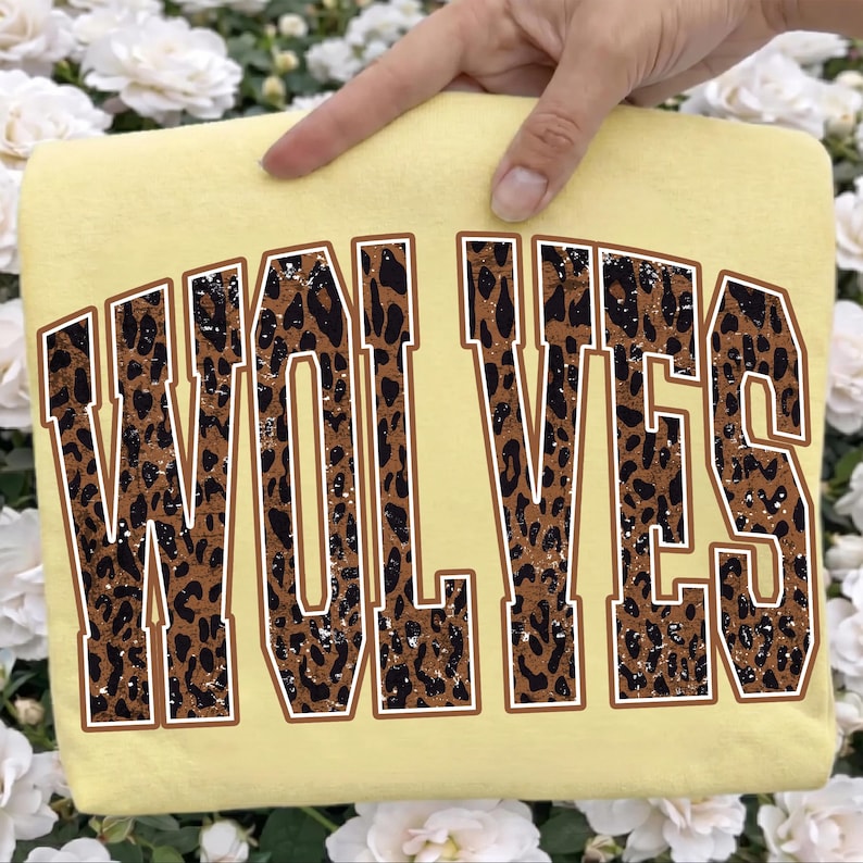 K&ouml;nnte beinhalten: Ein blassgelbes T-Shirt mit dem Wort WOLVES in gro&szlig;en Blockbuchstaben im College-Stil. Die Buchstaben haben ein braun-schwarzes Leopardenmuster mit einer abgenutzten, gesprenkelten Textur und einer d&uuml;nnen wei&szlig;en Umrandung um jedes Zeichen.