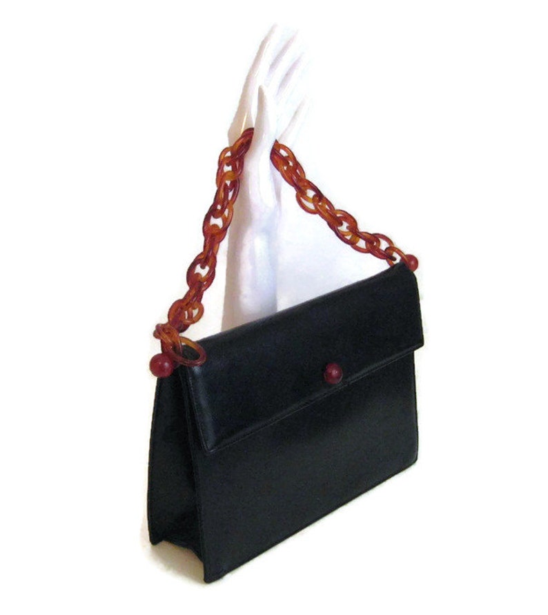 May include: A black leather handbag with a flap closure and a long, brown plastic chain strap. The strap has red plastic beads at the ends.