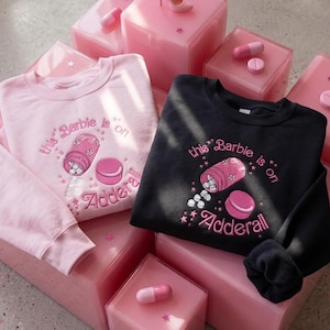 May include: Two sweatshirts, one light pink and one black, with the embroidered text "this Barbie is on Adderall". The design includes a pill bottle and a pink pill. The sweatshirts are displayed on pink translucent blocks.
