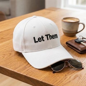 Puede incluir: Una gorra de b&eacute;isbol blanca con el texto Let Them bordado en hilo negro en el panel frontal. La gorra presenta una corona estructurada, una visera curva y ojales cosidos para la ventilaci&oacute;n, con un dise&ntilde;o limpio y sencillo.