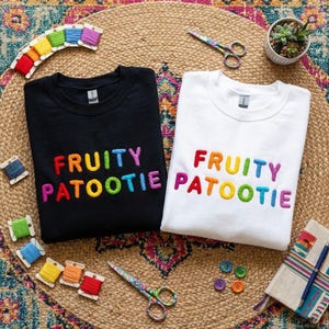 May include: A black sweatshirt and a white sweatshirt, each featuring the text "FRUITY PATOOTIE" embroidered in multicolored thread. The sweatshirts are surrounded by colorful embroidery floss spools, two pairs of scissors, buttons, and a notebook.