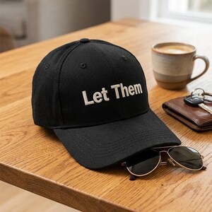 Puede incluir: Una gorra de b&eacute;isbol negra con el texto Let Them bordado en blanco en el panel frontal. La gorra cuenta con una corona estructurada, una visera curva y ojales cosidos para la ventilaci&oacute;n, presentando un dise&ntilde;o limpio y sencillo.
