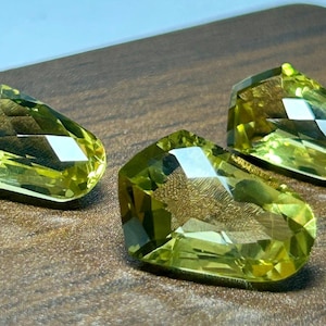 May include: Three faceted, light yellow-green gemstones with a pear-like shape are displayed on a wooden surface. The gems have multiple facets that reflect light, creating a sparkling effect. The background is a dark brown wood.