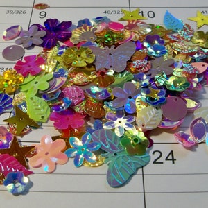 May include: A collection of iridescent sequins in various shapes, including flowers, butterflies, leaves, and stars. The sequins are in shades of pink, purple, green, and gold, scattered on a white grid background. These are ideal for craft projects.