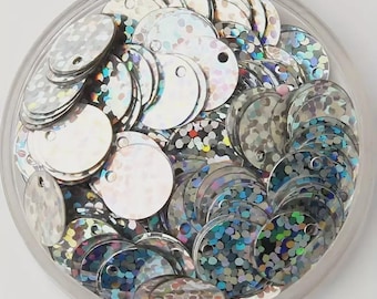May include: A clear plastic container filled with silver, iridescent sequins. The sequins are round with a small hole at the top for stringing. The sequins have a shiny, reflective surface, creating a sparkling effect.