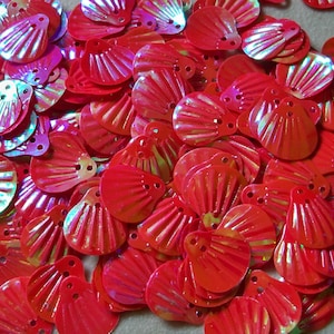 May include: A large pile of red iridescent sequins shaped like seashells. The sequins have a shiny, holographic finish.
