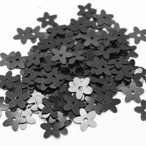 May include: A pile of black flower-shaped sequins. The sequins are about 1 cm in diameter and have a hole in the center.