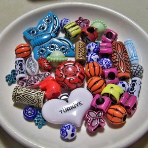 May include: A white ceramic dish filled with colorful plastic beads, including a blue butterfly bead, a white heart bead with the word "Turniye" written on it, and a variety of other shapes and designs.