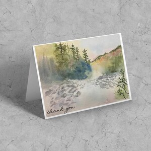 May include: Watercolor thank you card featuring a landscape scene with a river, trees, and mountains. The card has a white border and the words "thank you" are written in cursive at the bottom.
