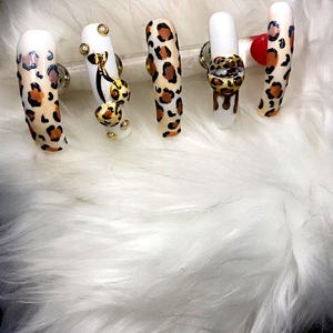 May include: A set of five long, artificial nails. The nails feature a white base with a leopard print design in brown and black. Some nails have gold and jewel accents. The nails are displayed on a white, fluffy surface.