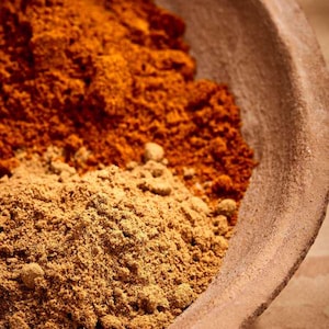 Puede incluir: Primer plano de un cuenco de madera lleno de dos tipos de especias. La mitad superior es de un rojo anaranjado vibrante, mientras que la mitad inferior es de un marrón claro, tostado. El borde del cuenco es de un marrón cálido y terroso.
