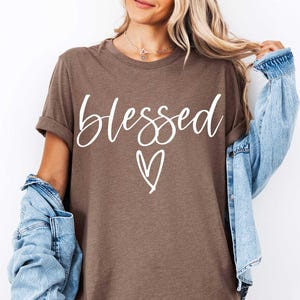 May include: Heather brown t-shirt with the word "blessed" in white script and a white heart graphic. The t-shirt has rolled-up sleeves and is worn with a light blue denim jacket.