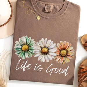 May include: A brown t-shirt with a daisy floral design in shades of green, white, and orange. The text "Life is Good" is printed in white script below the flowers. A gold necklace and a hat are next to the shirt.
