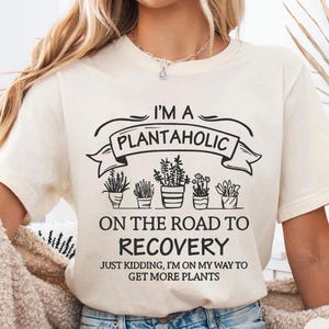 Op de afbeelding: Cr&egrave;mekleurig T-shirt met de zwarte tekst "I'M A PLANTAHOLIC ON THE ROAD TO RECOVERY". Daaronder illustraties van potplanten. Het shirt zegt ook "JUST KIDDING, I'M ON MY WAY TO GET MORE PLANTS."