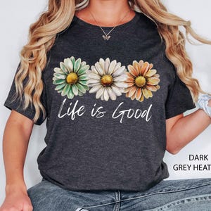 K&ouml;nnte beinhalten: Dunkelgraues Heather-T-Shirt mit drei G&auml;nsebl&uuml;mchen in Gr&uuml;n, Wei&szlig; und Orange mit gelben Zentren. Die Worte "Life is Good" sind in wei&szlig;er Schrift darunter gedruckt. Ein l&auml;ssiges Kleidungsst&uuml;ck f&uuml;r den Alltag.