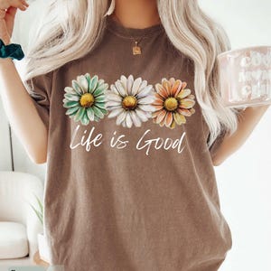 May include: Brown t-shirt featuring three daisy flowers in green, white, and orange with yellow centers. The words "Life is Good" are written in white script below the flowers. The shirt has a vintage, worn look.