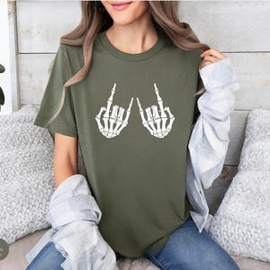 May include: A military green short-sleeve t-shirt featuring a white graphic print of two skeleton hands making the rock and roll hand sign. The shirt has a crew neckline and a standard fit, showcasing a bold skeletal design on the chest area.
