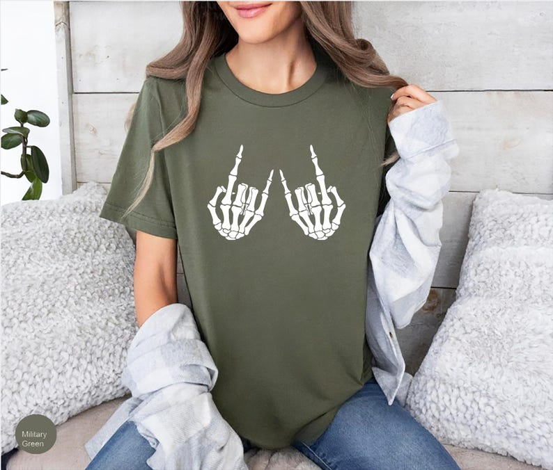 May include: A military green short-sleeve t-shirt featuring a white graphic print of two skeleton hands making the rock and roll hand sign. The shirt has a crew neckline and a standard fit, showcasing a bold skeletal design on the chest area.