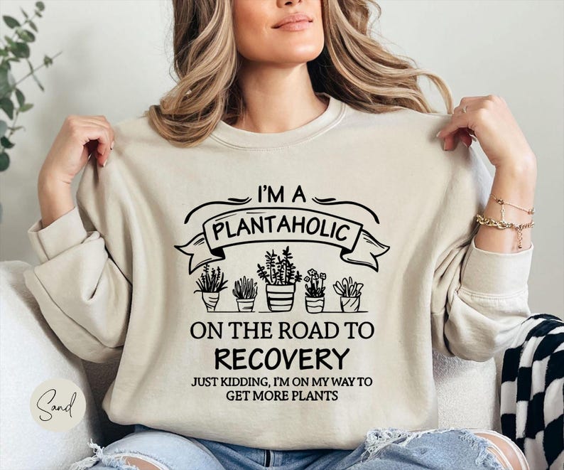 Op de afbeelding: Zandkleurige sweatshirt met de zwarte tekst "I'M A PLANTAHOLIC ON THE ROAD TO RECOVERY JUST KIDDING, I'M ON MY WAY TO GET MORE PLANTS". De afbeelding bevat illustraties van potplanten. De sweatshirt heeft een neutrale kleur.