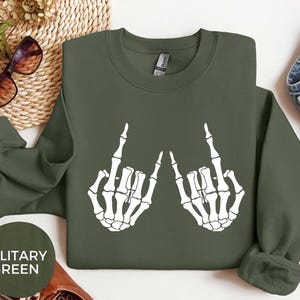 May include: A military green long-sleeve sweatshirt featuring a white graphic print of two skeleton hands making a rock and roll hand gesture. The sweatshirt has a crew neckline and long sleeves with ribbed cuffs, displayed flat with a circular label.