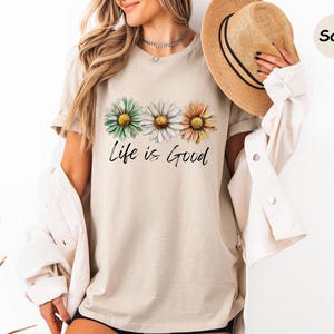 K&ouml;nnte beinhalten: Sandfarbenes T-Shirt mit drei Aquarell-G&auml;nsebl&uuml;mchen in Gr&uuml;n, Wei&szlig; und Orange. Der Schriftzug "Life is Good" steht in zarter Schrift unter den Blumen. Das Shirt hat kurze &Auml;rmel und einen Rundhalsausschnitt.