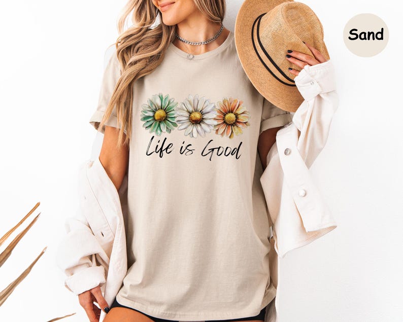 K&ouml;nnte beinhalten: Sandfarbenes T-Shirt mit drei Aquarell-G&auml;nsebl&uuml;mchen in Gr&uuml;n, Wei&szlig; und Orange. Der Schriftzug "Life is Good" steht in zarter Schrift unter den Blumen. Das Shirt hat kurze &Auml;rmel und einen Rundhalsausschnitt.