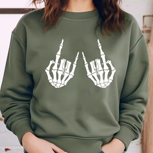 May include: An olive green crewneck sweatshirt featuring a white graphic print of two skeletal hands positioned with the index and pinky fingers extended in a rock and roll gesture. The long-sleeved garment has a ribbed collar, cuffs, and hem.