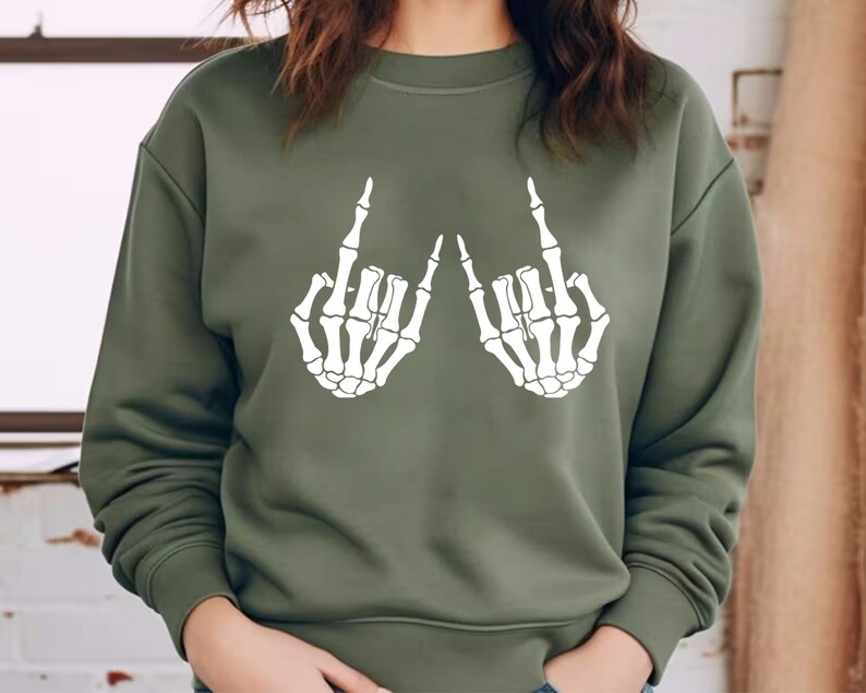 May include: An olive green crewneck sweatshirt featuring a white graphic print of two skeletal hands positioned with the index and pinky fingers extended in a rock and roll gesture. The long-sleeved garment has a ribbed collar, cuffs, and hem.