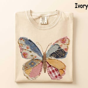 May include: Ivory-colored t-shirt featuring a colorful butterfly applique. The butterfly design incorporates various fabric patterns, including floral, denim, and plaid. The shirt has a crew neck and short sleeves. The tag reads "Comfort Colors".