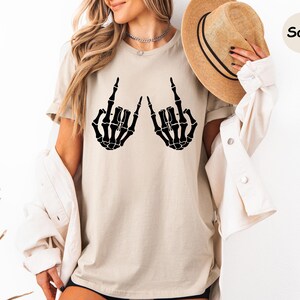 May include: A sand-colored t-shirt featuring a black graphic of two skeleton hands making the rock and roll sign. The shirt is paired with a light-colored button-down shirt and a tan fedora hat with a black band. The text Sand is visible.