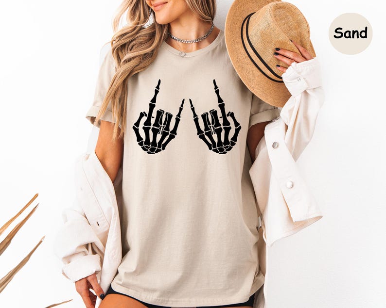 May include: A sand-colored t-shirt featuring a black graphic of two skeleton hands making the rock and roll sign. The shirt is paired with a light-colored button-down shirt and a tan fedora hat with a black band. The text Sand is visible.