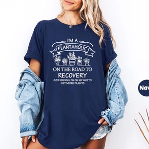 Op de afbeelding: Marineblauwe t-shirt met witte tekst: "I'M A PLANTAHOLIC ON THE ROAD TO RECOVERY". Het shirt zegt ook "JUST KIDDING, I'M ON MY WAY TO GET MORE PLANTS". Het t-shirt heeft een ronde hals en korte mouwen.