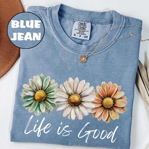 May include: A blue jean Comfort Colors t-shirt with three daisy flowers in green, white, and orange. The words "Life is Good" are printed in white script across the bottom. A gold necklace with a small pendant is draped across the shirt.