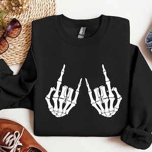 May include: A black long-sleeve crewneck sweatshirt featuring a white graphic print of two skeleton hands positioned to display the rock and roll hand sign. The sweatshirt has a ribbed collar, cuffs, and hem, with a standard fit and solid black fabric.
