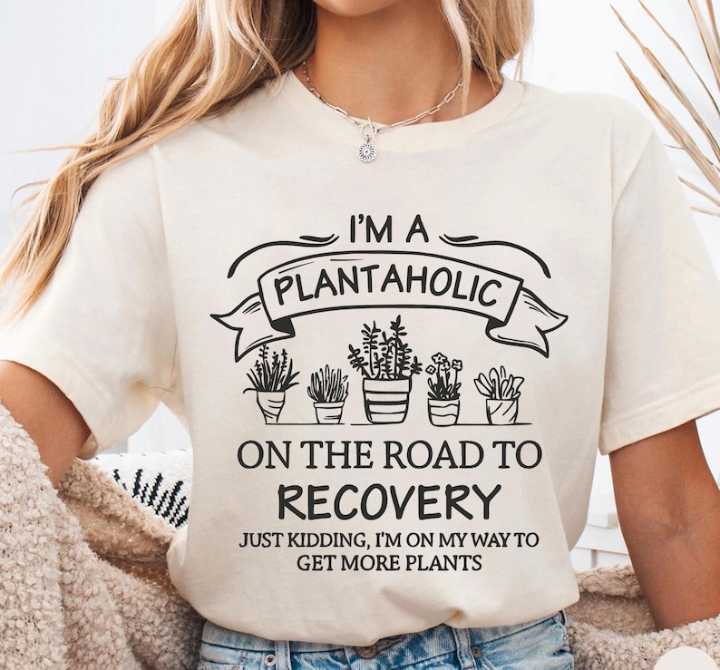 Op de afbeelding: Cr&egrave;mekleurig T-shirt met de zwarte tekst "I'M A PLANTAHOLIC ON THE ROAD TO RECOVERY". Daaronder illustraties van potplanten. Het shirt zegt ook "JUST KIDDING, I'M ON MY WAY TO GET MORE PLANTS."