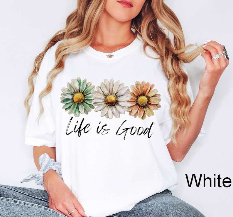 K&ouml;nnte beinhalten: Wei&szlig;es T-Shirt mit drei G&auml;nsebl&uuml;mchen in Gr&uuml;n, Wei&szlig; und Orange mit gelben Zentren. Der Schriftzug "Life is Good" ist in schwarzer Schrift geschrieben. Das T-Shirt hat einen l&auml;ssigen Stil.