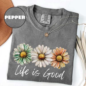 May include: A gray t-shirt with a daisy floral design in green, white, and orange. The words "Life is Good" are printed in white script below the flowers. A silver necklace with a small pendant is draped on the shirt.