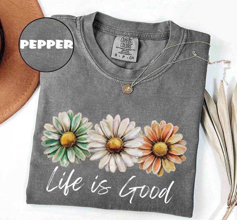 May include: A gray t-shirt with a daisy floral design in green, white, and orange. The words "Life is Good" are printed in white script below the flowers. A silver necklace with a small pendant is draped on the shirt.