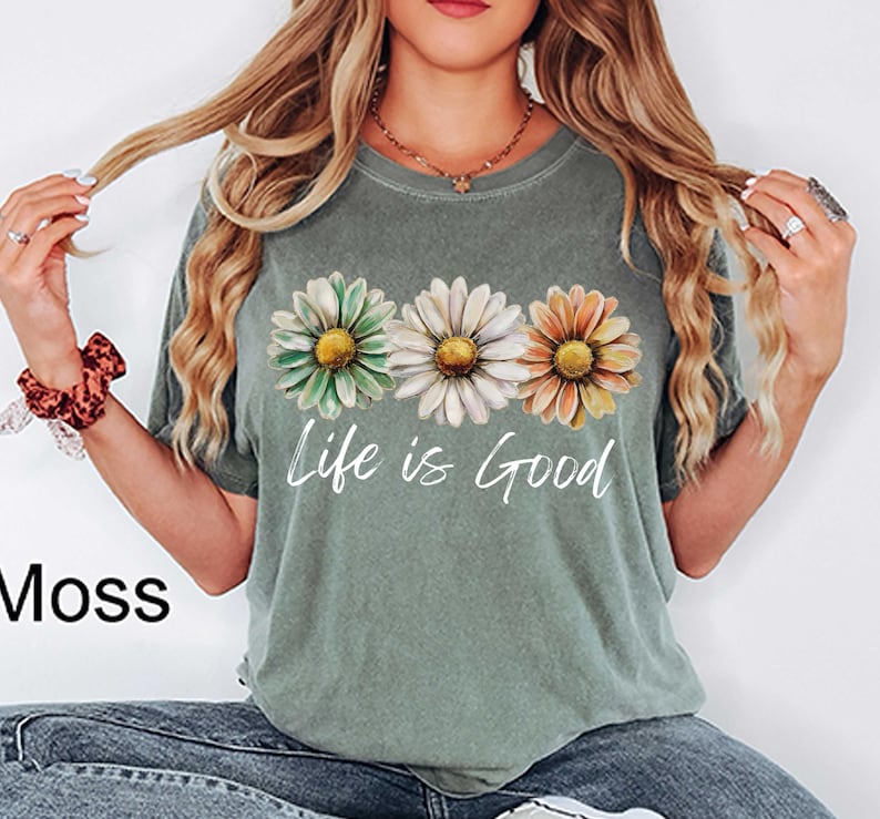 May include: Moss green t-shirt featuring three daisy flowers in green, white, and orange. The words "Life is Good" are printed in white script below the flowers. The shirt is a casual style.