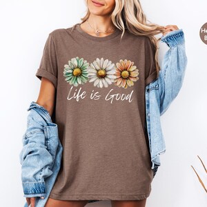K&ouml;nnte beinhalten: Heatherbraunes T-Shirt mit drei G&auml;nsebl&uuml;mchen in Gr&uuml;n, Wei&szlig; und Orange mit gelben Zentren. Die Worte "Life is Good" sind in einer kursiven Schrift unter den Blumen geschrieben. Das Shirt hat hochgekrempelte &Auml;rmel.