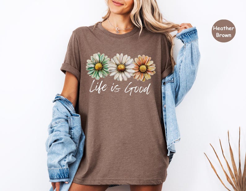 K&ouml;nnte beinhalten: Heatherbraunes T-Shirt mit drei G&auml;nsebl&uuml;mchen in Gr&uuml;n, Wei&szlig; und Orange mit gelben Zentren. Die Worte "Life is Good" sind in einer kursiven Schrift unter den Blumen geschrieben. Das Shirt hat hochgekrempelte &Auml;rmel.